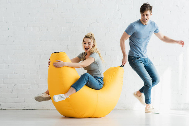 handsome man pulling yellow bean bag chair with cheerful woman  - Fotografie, Obrázek