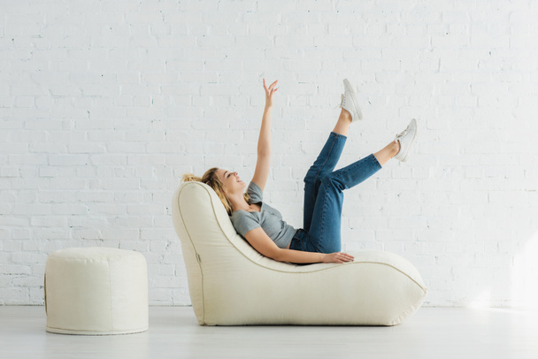 cheerful blonde girl lying on  bean bag chair and gesturing near brick wall  - Foto, immagini