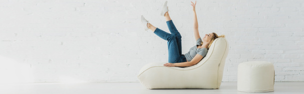 panoramic shot of cheerful blonde girl lying on  bean bag chair and gesturing near brick wall  - Фото, изображение