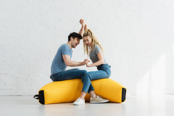 pretty blonde girl and handsome man holding hands while sitting on yellow bean bag chair - Photo, Image