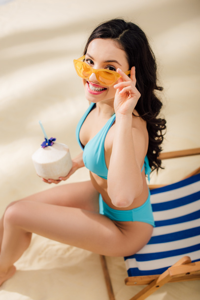 beautiful smiling girl in bikini with coconut cocktail sitting on deck chair and looking at camera on beach - Photo, Image