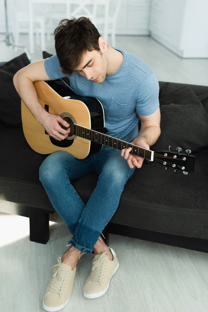 jóképű zenész játszik akusztikus gitár ülve keresztbe lábak a kanapén   - Fotó, kép