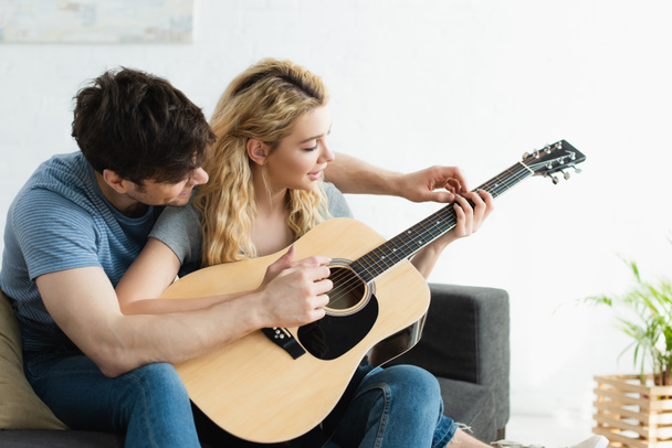 handsome man teaching blonde happy woman playing acoustic guitar at home  - Foto, immagini