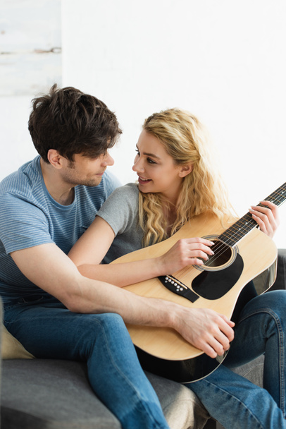 happy blonde young woman holding acoustic guitar near cheerful man sitting on sofa  - Fotografie, Obrázek