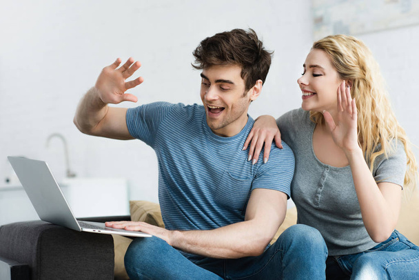 cheerful man and happy blonde girl waving hands while having video call on laptop  - 写真・画像