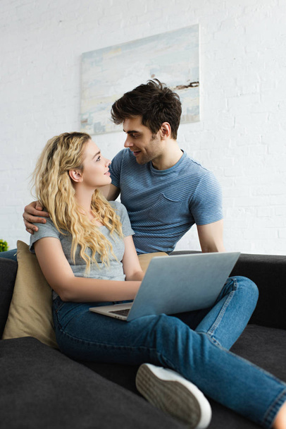 schöner Mann umarmt und schaut glückliches blondes Mädchen mit Laptop an  - Foto, Bild
