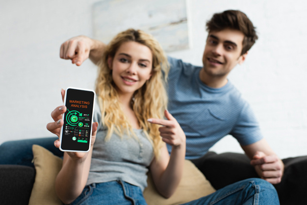 selective focus of happy man and blonde woman pointing with fingers at smartphone with marketing analysis lettering on screen - Foto, Imagem