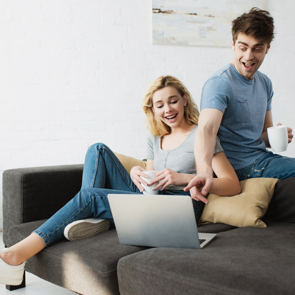 happy man holding cup and pointing with finger at laptop near cheerful blonde girl sitting on sofa  - Photo, image