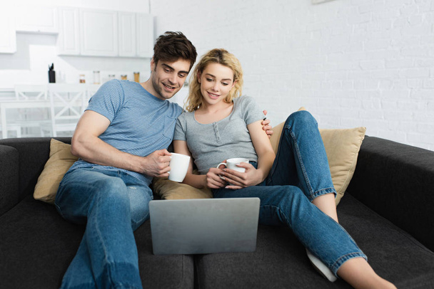 happy man holding cup and watching movie with attractive girl on laptop  - Fotografie, Obrázek