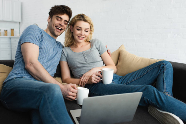 happy man holding cup and watching movie with cheerful girl on laptop  - Фото, зображення