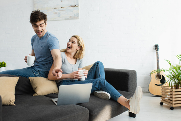 happy man pointing with finger at laptop while holding cup near attractive blonde girl  - Фото, зображення