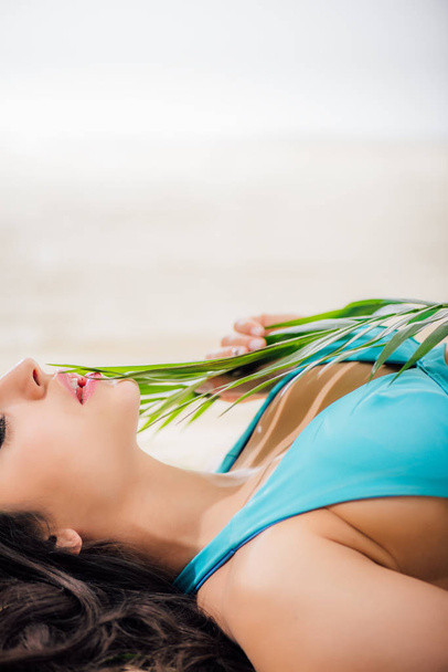 cropped view of sexy girl in bikini with eyes closed posing with green leaf while lying on beach with copy space - Foto, immagini