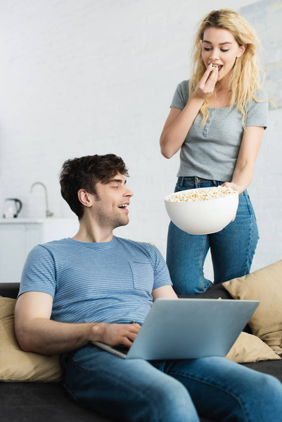 happy blonde girl holding bowl with popcorn near man using laptop  - Zdjęcie, obraz