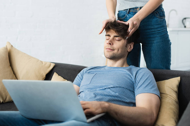 cropped view of woman touching head of happy man sitting on sofa with laptop - 写真・画像