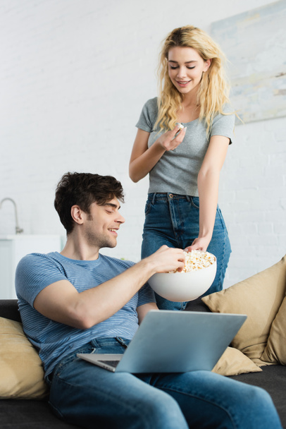 szczęśliwy blond dziewczyna trzyma miskę z popcorn w pobliżu wesoły człowiek siedzi z laptopem na kanapie - Zdjęcie, obraz