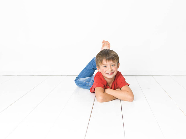 cute blond boy in red t-shirt and blue jeans lying with crossed arms on white wooden floor in front of white background - Zdjęcie, obraz