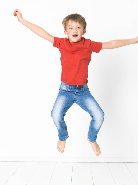 cute blond boy in red t-shirt and blue jeans jumping on white wooden floor in front of white background - Photo, image