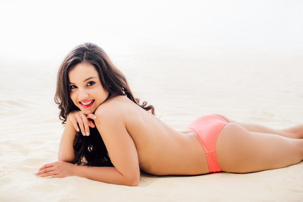 hermosa mujer joven en topless mirando a la cámara, sonriendo y relajándose en la playa de arena
 - Foto, imagen