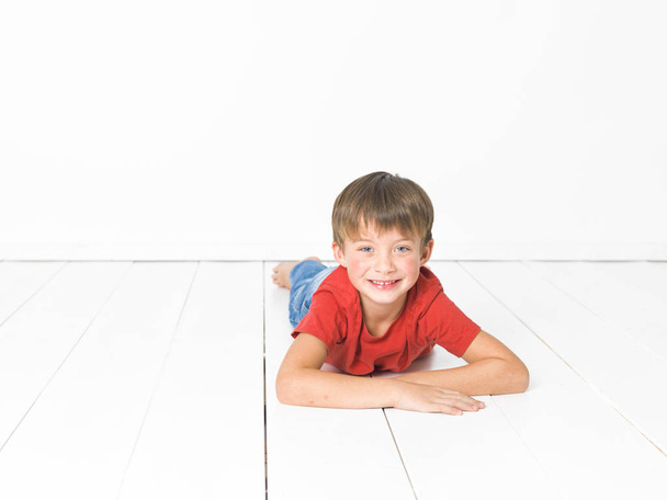 cute blond boy in red t-shirt and blue jeans lying on white wooden floor in front of white background - Valokuva, kuva
