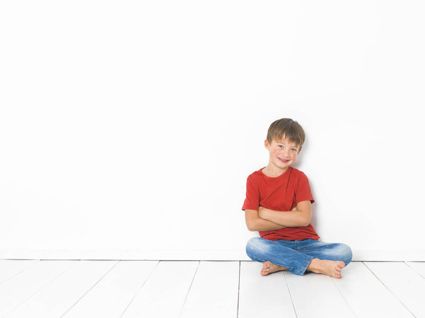 cute blond boy in red t-shirt and blue jeans sitting with crossed arms on white wooden floor in front of white background - Foto, imagen