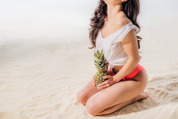 cropped view of girl sitting and posing with pineapple on sandy beach - Fotografie, Obrázek