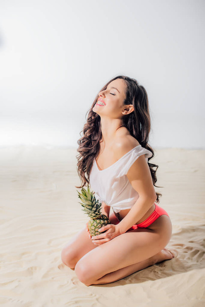 красива усміхнена дівчина сидить і позує з ананасом на піщаному пляжі
 - Фото, зображення
