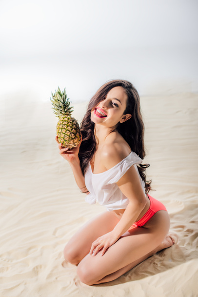 mooi glimlachend meisje zittend, kijkend naar camera en poseren met ananas op zandstrand - Foto, afbeelding