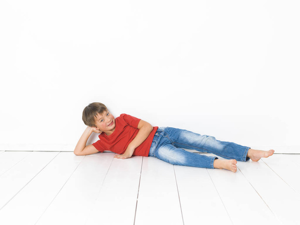 cute blond boy in red t-shirt and blue jeans lying on white wooden floor in front of white background - Fotoğraf, Görsel