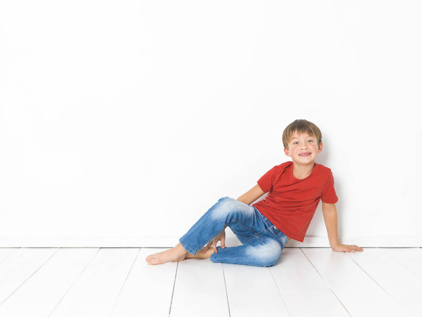 cute blond boy in red t-shirt and blue jeans sitting on white wooden floor in front of white background - Foto, Imagem