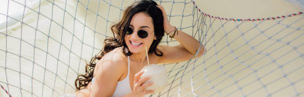 panoramic shot of beautiful smiling girl in sunglasses with coconut cocktail lying in hammock on beach - Photo, Image