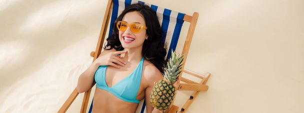 panoramic shot of beautiful smiling girl in bikini and sunglasses with pineapple lying on deck chair on beach - Photo, image