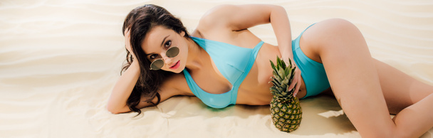 panoramic shot of beautiful girl in bikini and sunglasses with pineapple Looking At Camera while lying on beach - Photo, Image