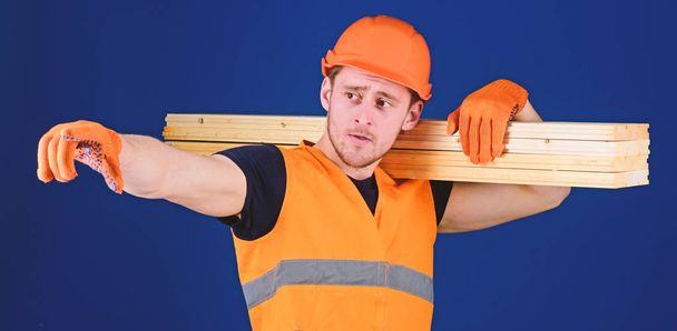 Man in helmet, hard hat and protective gloves pointing direction, blue background. Wooden materials concept. Carpenter, woodworker, strong builder on thoughtful face carries wooden beam on shoulder - Foto, Bild