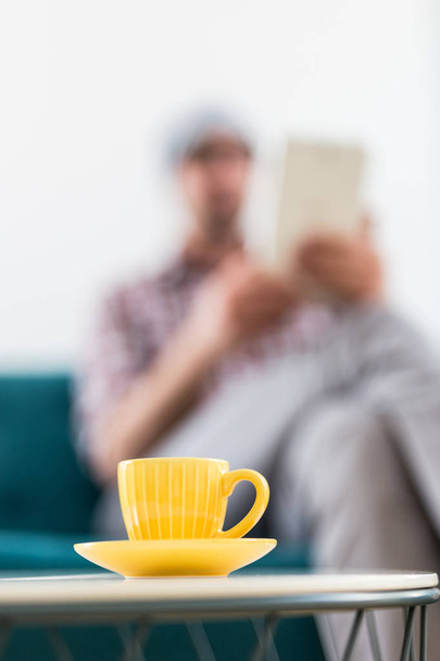 Yellow cup of coffee on small table and fashionable businessman with digital tablet in the background. - Foto, imagen