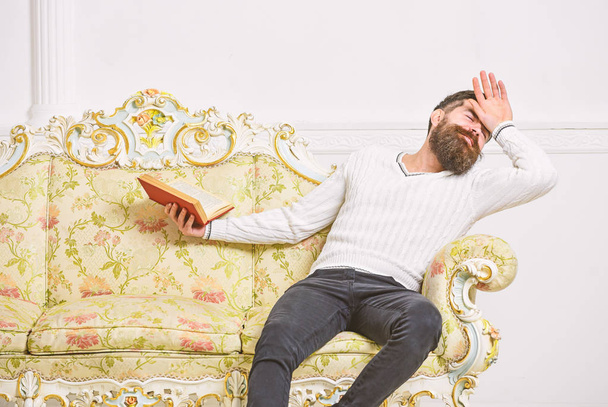 Macho on dramatic empathizing face reading book. Empathy concept. Guy reading book with empathy. Man with beard and mustache sits on baroque style sofa, holds book, white wall background - Photo, image
