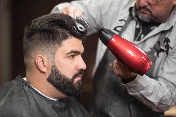 Κλείνω πάνω θέα του master barber κάνει χτένισμα με στεγνωτήρα μαλλιών στο κομμωτήριο. - Φωτογραφία, εικόνα