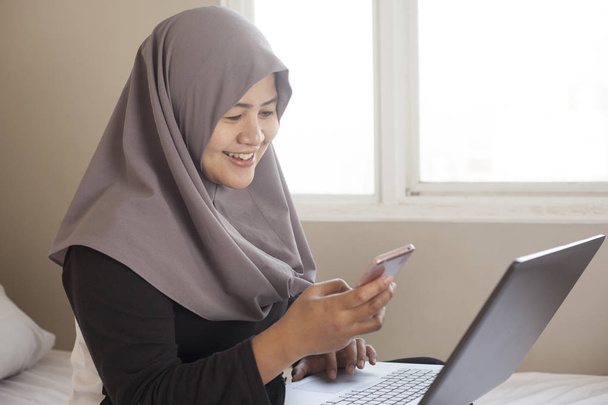 Mulher muçulmana feliz trabalhando com laptop e telefone inteligente em seu ser
 - Foto, Imagem
