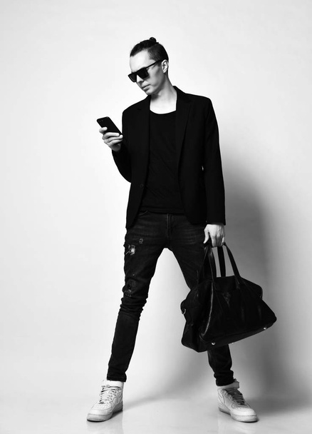 Young modern man with a bun on head in sunglasses and with a big bag is typing a message or read news on a cell phone - Foto, Imagem
