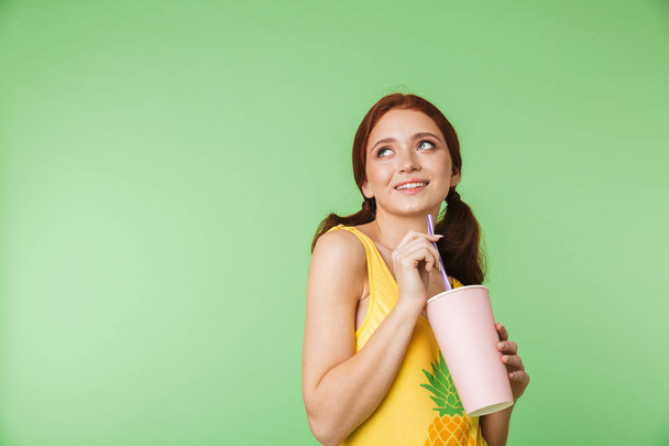 Beautiful happy young redhead girl posing isolated over green wall background drinking cocktail. - Photo, Image