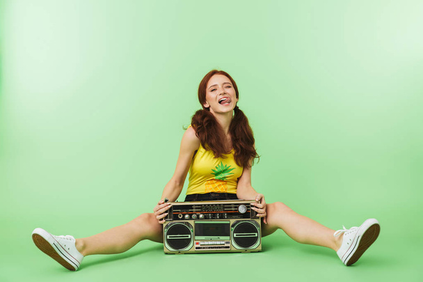 Happy young redhead girl posing isolated over green wall background with boombox. - Fotoğraf, Görsel