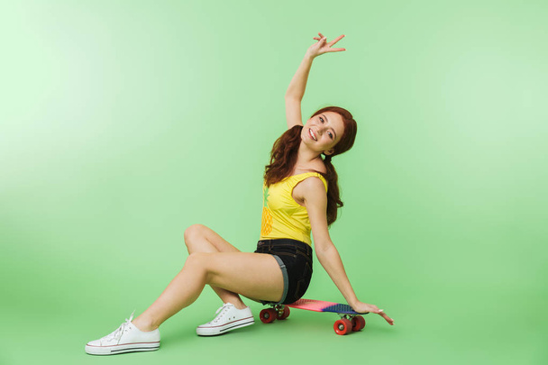 Happy young redhead girl posing isolated over green wall background with skateboard. - Foto, afbeelding