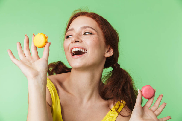 Redhead girl posing isolated over green wall background holding sweeties macaroons. - Fotoğraf, Görsel