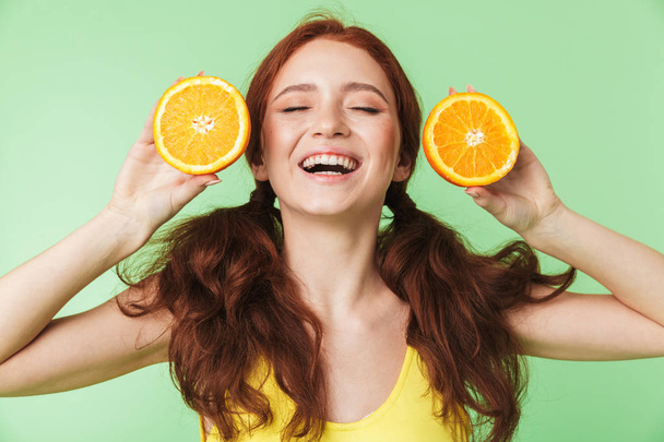 Young redhead girl posing isolated over green wall background with citrus vitamins fruits. - Фото, изображение