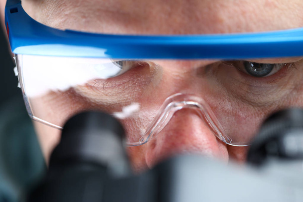 Male laboratory worker eyes looking at microscope wearing goggles - Photo, Image