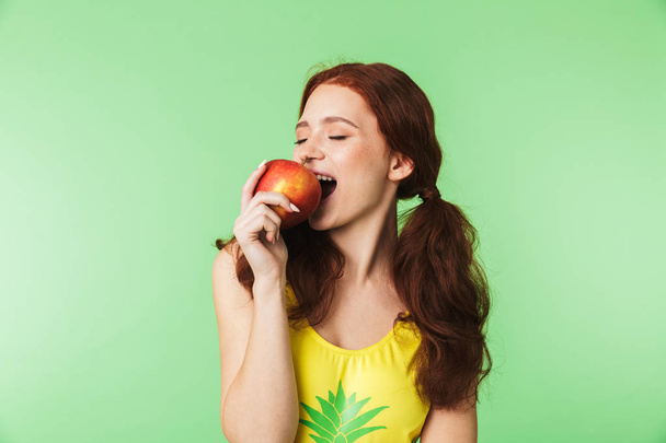 Emotional young redhead girl posing isolated over green wall background with apple. - Фото, изображение