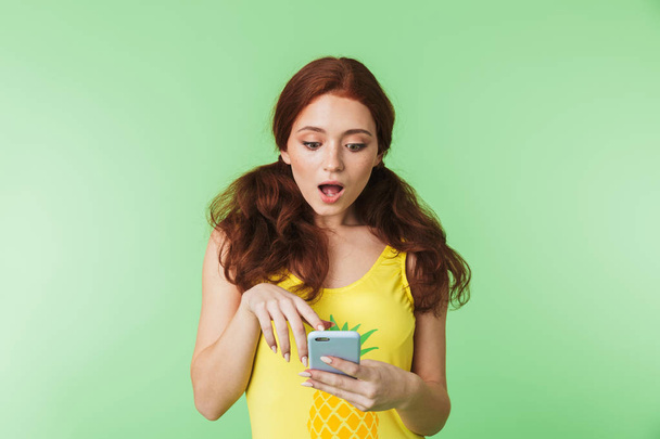 Beautiful shocked emotional young redhead girl posing isolated over green wall background using mobile phone. - Foto, afbeelding