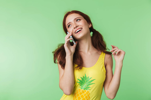 Beautiful young redhead girl posing isolated over green wall background talking by mobile phone. - Фото, зображення