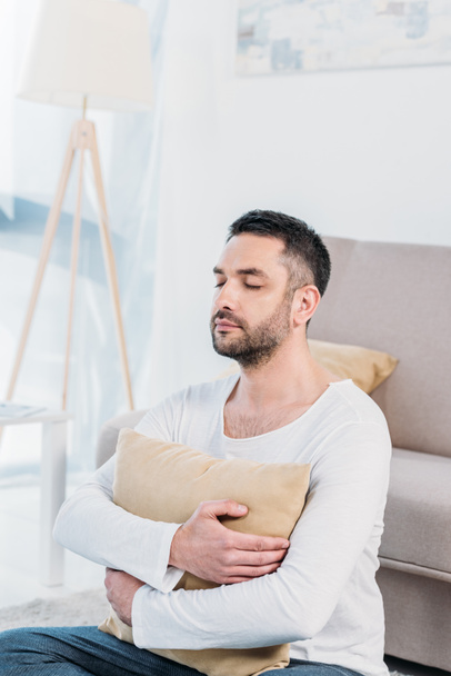 handsome man with eyes closed sitting on carpet and hugging pillow at home - Photo, Image