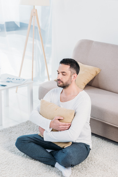 handsome man with eyes closed sitting on carpet and hugging pillow at home in living room - Fotografie, Obrázek
