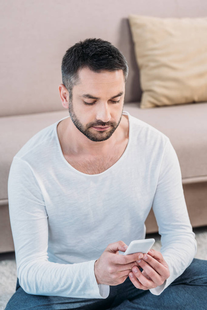 focused handsome man in casual clothes sitting and using smartphone at home - Fotografie, Obrázek